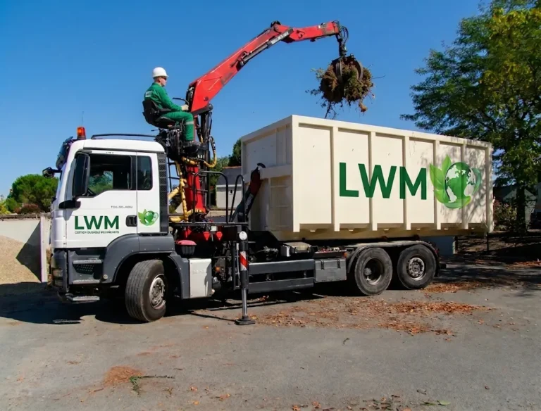 Camion de collecte de déchets LWM « Gestion durable des déchets » équipé d’une grue hydraulique rouge. Un opérateur charge un ballot de matières dans un grand conteneur blanc LWM en extérieur sous un ciel bleu ensoleillé.